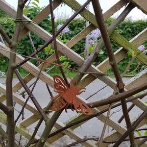 K&ouml;nnte beinhalten: Ein rostiges Metall-Bienen-Ornament, das an einem Holzgitter mit gr&uuml;nem Laub und lila Bl&uuml;ten im Hintergrund h&auml;ngt.