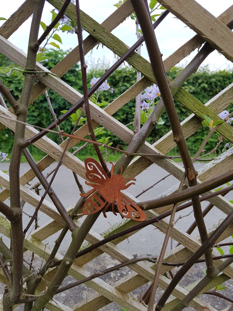 K&ouml;nnte beinhalten: Ein rostiges Metall-Bienen-Ornament, das an einem Holzgitter mit gr&uuml;nem Laub und lila Bl&uuml;ten im Hintergrund h&auml;ngt.