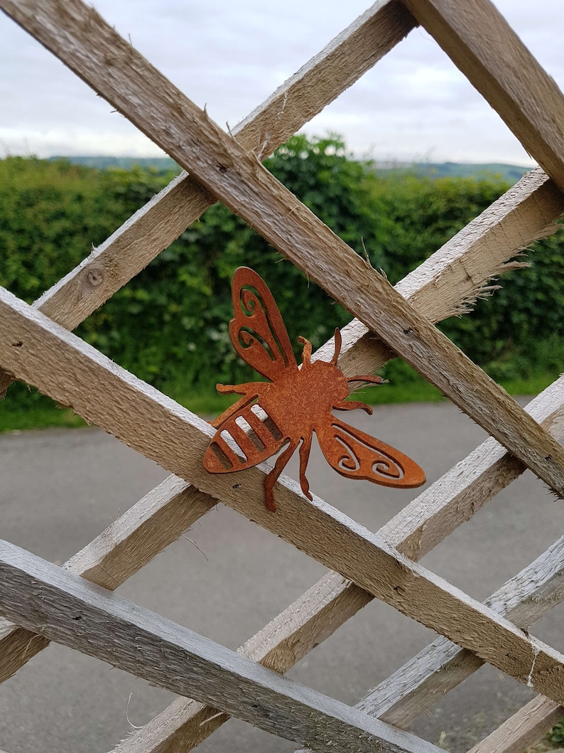 K&ouml;nnte beinhalten: Ein rostiger brauner Metallausschnitt einer Biene, der an einem Holzlattenzaun befestigt ist. Die Biene hat einen gestreiften K&ouml;rper und detaillierte Fl&uuml;gel.