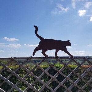 May include: A black metal silhouette of a cat walking on a white wooden fence with a diamond lattice pattern. The fence is in front of a green field and a blue sky with white clouds.