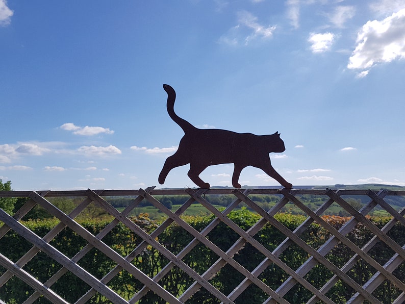 May include: A black metal silhouette of a cat walking on a white wooden fence with a diamond lattice pattern. The fence is in front of a green field and a blue sky with white clouds.