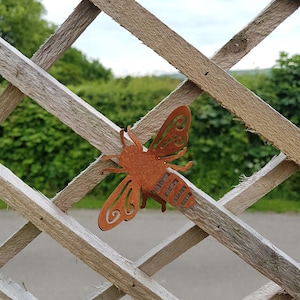 K&ouml;nnte beinhalten: Ein rostiges braunes Metall-Bienen-Ausschnitt mit einem dekorativen Design. Die Biene h&auml;ngt an einem Holzgitterzaun.