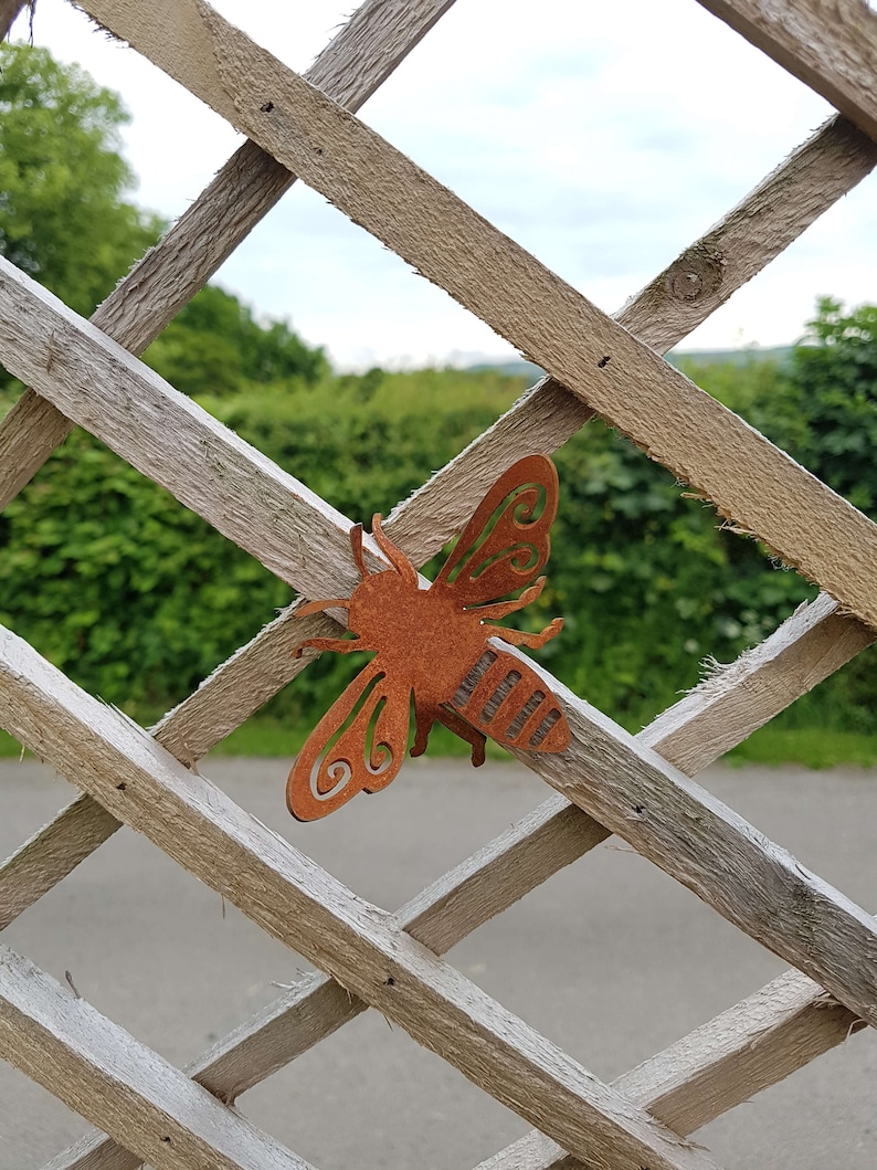 K&ouml;nnte beinhalten: Ein rostiges braunes Metall-Bienen-Ausschnitt mit einem dekorativen Design. Die Biene h&auml;ngt an einem Holzgitterzaun.
