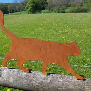 May include: A rusty metal silhouette of a cat walking along a wooden fence post. The cat is facing to the right and has a long tail. The background is a green field with a few trees in the distance.