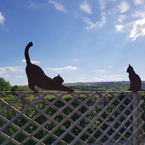 May include: Two black metal cat silhouettes on a wooden fence against a blue sky with white clouds. The cat on the left is stretching, while the cat on the right is sitting.