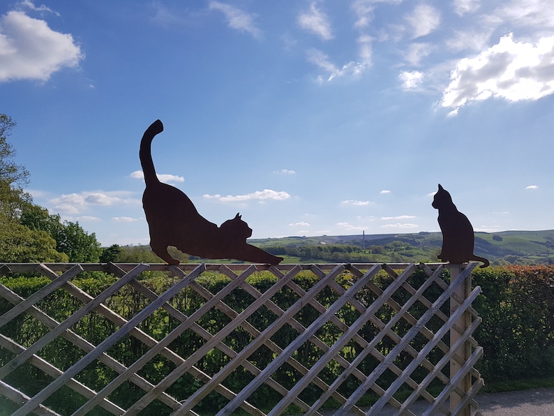 May include: Two black metal cat silhouettes on a wooden fence against a blue sky with white clouds. The cat on the left is stretching, while the cat on the right is sitting.