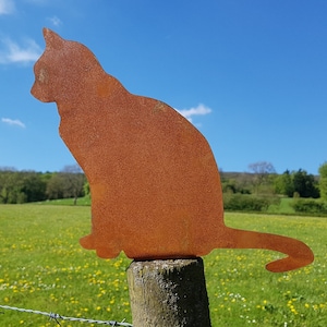 May include: A rusty metal silhouette of a cat sitting on a wooden post. The cat is facing to the left and has a long tail. The background is a green field with yellow flowers.