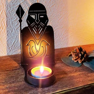 Pode incluir: Um castiçal de metal preto em forma de guerreiro viking com uma lança. Há uma vela de chá acesa dentro, emitindo um brilho quente. O castiçal está sobre uma superfície de madeira. Aproximadamente 15 cm de altura.