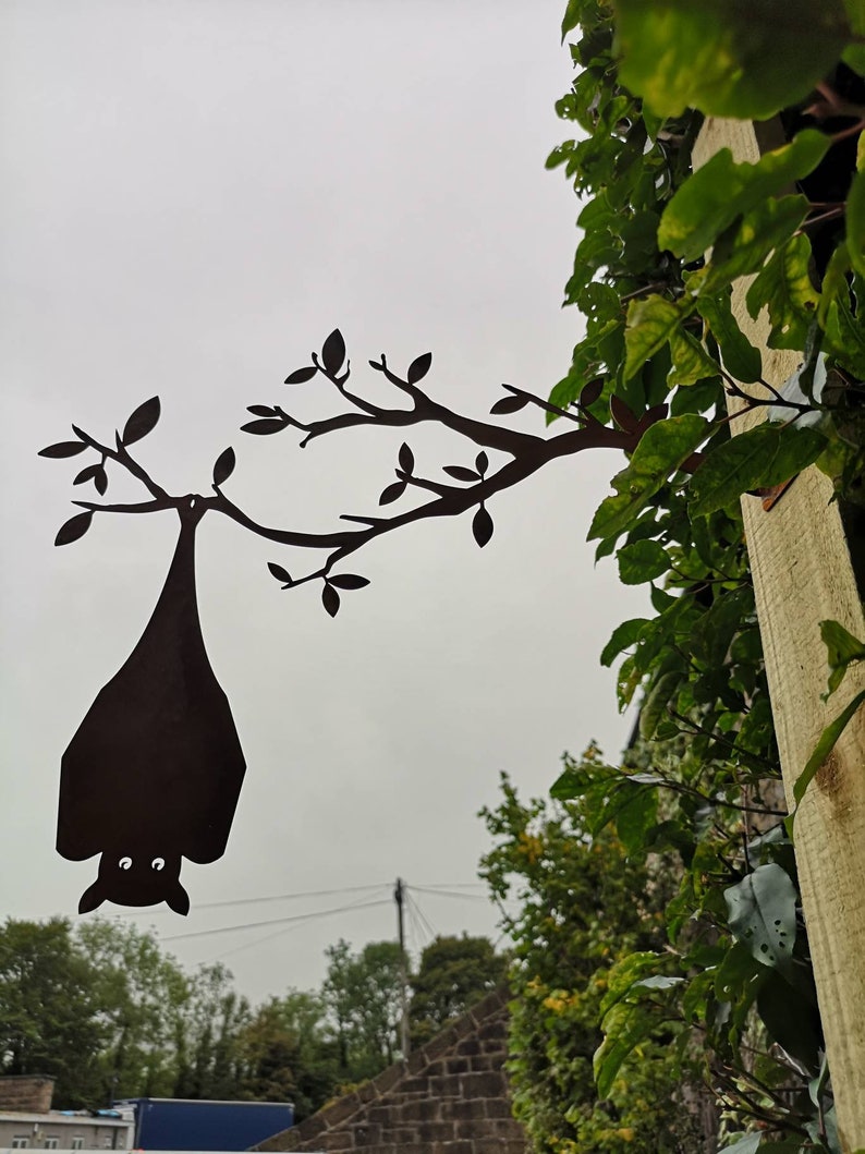 May include: A black metal silhouette of a bat hanging upside down from a tree branch. The silhouette is attached to a wooden post.