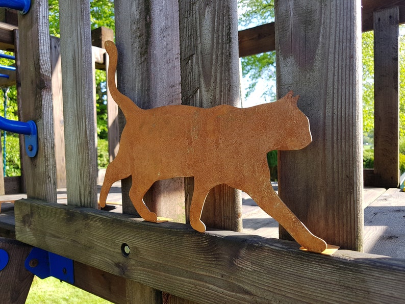 May include: A rusty metal cat silhouette, mounted on a wooden fence post. The cat is facing right and has a long tail.