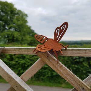 K&ouml;nnte beinhalten: Eine rostige Metall-Bienenskulptur mit filigranen Details, die auf einem Holzzaun vor einem verschwommenen Hintergrund aus gr&uuml;nen B&auml;umen und einem bew&ouml;lkten Himmel sitzt.