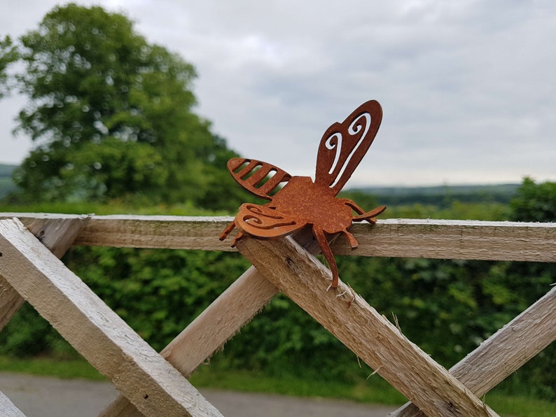 K&ouml;nnte beinhalten: Eine rostige Metall-Bienenskulptur mit filigranen Details, die auf einem Holzzaun vor einem verschwommenen Hintergrund aus gr&uuml;nen B&auml;umen und einem bew&ouml;lkten Himmel sitzt.