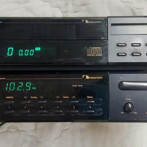 以下が含まれることがあります： 2つの黒いNakamichiオーディオコンポーネント：CDプレーヤーとAM/FMステレオレシーバー。CDプレーヤーは緑色で「0 0.00」を表示します。レシーバーは緑色で「102.9」を表示します。どちらにもさまざまなボタンとコントロールがあります。