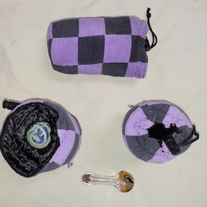 May include: A collection of corduroy pouches and a glass pipe. The pouches are in shades of purple and gray, with a checkerboard pattern on one and drawstring closures. The glass pipe has a clear stem and colorful accents.