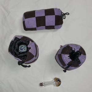 May include: A collection of corduroy pouches in a checkerboard pattern of brown and lavender. One pouch is rectangular, and two are round with drawstrings. A clear glass pipe with brown and gold accents is also present.