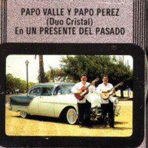 May include: Two men, dressed in casual clothing, stand in front of a vintage white and green car. They are holding acoustic guitars and smiling. The text "PAPO VALLE Y PAPO PEREZ (Duo Cristal) En UN PRESENTE DEL PASADO" is visible on the image.