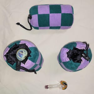 May include: A collection of corduroy pouches and a glass pipe. The pouches are in a checkerboard pattern of green and lavender. One pouch is rectangular, and two are round. The glass pipe is clear with yellow, orange, and black accents.