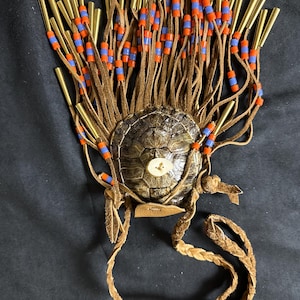 May include: A handcrafted turtle shell pendant with brass tubes, leather fringe, and a braided leather cord. The fringe is adorned with orange and blue beads. The pendant has a natural, earthy aesthetic.