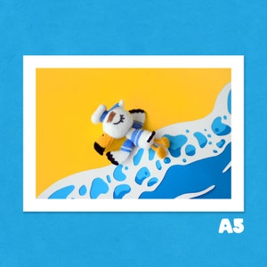 Peut inclure: Un jouet en peluche de mouette blanc et bleu avec un bec jaune et des yeux noirs est allongé sur une découpe en papier blanc et bleu d'une scène de plage. La mouette porte un chapeau de marin bleu et blanc à rayures.