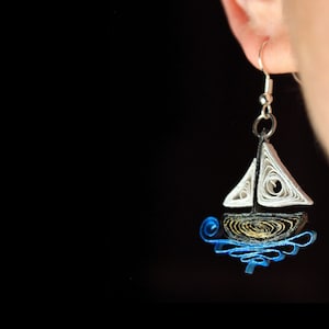 Peut inclure: Boucle d'oreille artisanale en forme de voilier. La voile blanche contraste avec la coque sombre, et des volutes bleues représentent l'eau. La boucle d'oreille, suspendue à un crochet argenté, se détache sur un fond noir. Fabriqué en papier roulé.