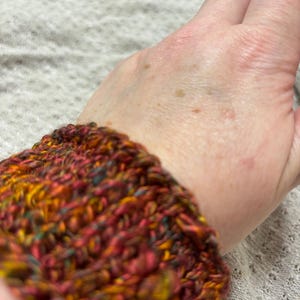 Könnte beinhalten: Nahaufnahme eines gehäkelten Armbands. Das Armband besteht aus Garn in Rot-, Orange-, Gelb- und Grüntönen. Die Textur des Garns ist sichtbar, mit einer leicht unebenen Oberfläche.