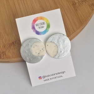 Pode incluir: Um par de brincos de meia lua prateados e brancos com textura salpicada. Os brincos são exibidos em um cartão com o texto "Holli Colors - Design" e "@hollicolorsdesign MADE IN PORTUGAL".