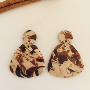 May include: A pair of handmade earrings with a marbled design in shades of brown, beige, and gold. The earrings have a circular top and a triangular bottom. Cinnamon sticks are in the background.
