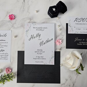 May include: Wedding invitation suite featuring a central invitation card with the names "Holly and Nathan", RSVP card, details card, and black envelopes. The suite includes a ring box, pearls, and floral accents, all arranged on a marble surface.