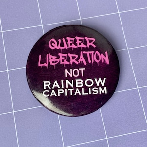 May include: A purple button with the text "Queer Liberation Not Rainbow Capitalism" in pink and white lettering.