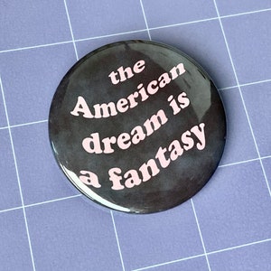 May include: A black button with the text "the American dream is a fantasy" in pink lettering.