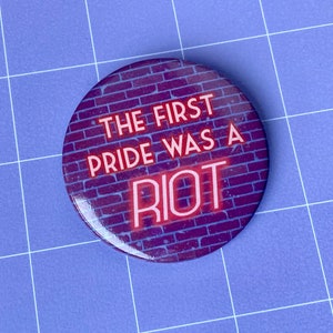 May include: A round button with a brick wall background and the text "THE FIRST PRIDE WAS A RIOT" in pink neon letters.