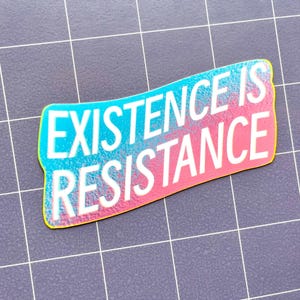 May include: A sticker with the words "EXISTENCE IS RESISTANCE" in white, with a gradient of blue and pink. The sticker has a yellow outline and is on a gray grid background.