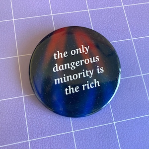 May include: Circular button with a glossy finish. The button features a dark blue and red background with the white text: "the only dangerous minority is the rich."