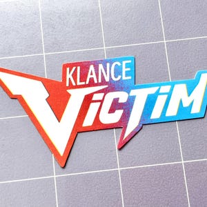May include: A die-cut sticker with the words "KLANCE Victim" in white, red, and blue. The word "KLANCE" is in a rectangular box, and "Victim" is in a stylized shape. The sticker is on a gray grid background.