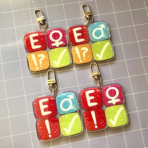 Puede incluir: Dos llaveros con colgantes cuadrados de colores. Cada colgante presenta una letra, un símbolo o un signo de puntuación. Los colgantes son rojos, naranjas, verdes y azules con llaveros dorados. Los llaveros tienen las letras "E", "Q" y los símbolos masculino y femenino.