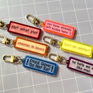 May include: Six rectangular acrylic keychains with gold-tone lobster clasps. Each features a unique phrase: plot what plot, you have already left kudos here :), enemies to lovers, hurt no comfort, Dead Dove: Do Not Eat!, and no beta we die like men.