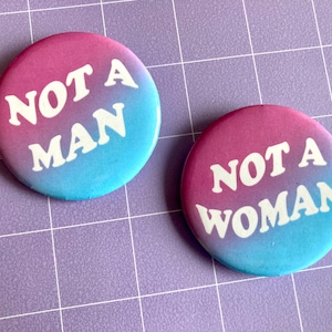 Puede incluir: Dos insignias redondas con un degradado de rosa y azul. Una dice "NOT A MAN", la otra "NOT A WOMAN" en texto blanco. El fondo presenta un degradado de color.