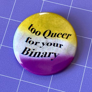 May include: A yellow, white, and purple button with the text "too queer for your binary" in black.