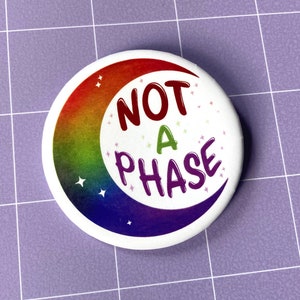 Puede incluir: Un botón blanco con una luna creciente arcoíris y el texto "NOT A PHASE" en letras de colores.