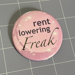 May include: A circular pin-back button with a light pink, glittery background. The text "rent lowering Freak" is printed in black across the center, with the word "rent" and "lowering" in a sans-serif font and "Freak" in a cursive script font.