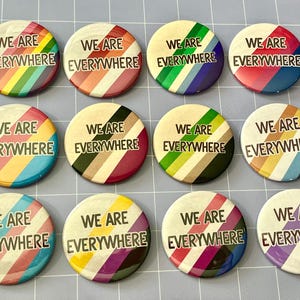 May include: Twelve circular pinback buttons arranged in a grid, each featuring diagonal stripes in various colour combinations and the black text WE ARE EVERYWHERE printed in the centre. The buttons display diverse pride flag colour patterns.