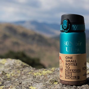 May include: A teal water bottle with a black lid and a brown paper label. The bottle has the text "80 Noir Ultra" printed on it. The label reads "One Small Bottle to Change the World."