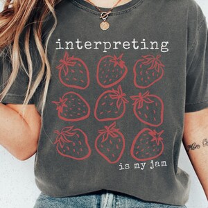 May include: Dark grey t-shirt with the text "interpreting is my jam" and a pattern of nine red strawberries. The shirt is a casual style, suitable for everyday wear.