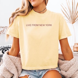 May include: A light yellow t-shirt with the text "LIVE FROM NEW YORK" in a simple font. The shirt is paired with denim shorts and a beige sweater. The overall aesthetic is casual and relaxed.
