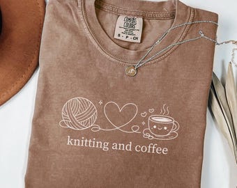 May include: A brown Comfort Colors t-shirt with a white graphic of a yarn ball, heart, and coffee cup, with the words "knitting and coffee" below. A silver necklace with a small pendant is draped across the shirt.