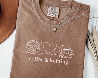 May include: A brown Comfort Colors t-shirt with a white graphic of a yarn ball, heart, and coffee cup with the text "coffee & knitting." A silver necklace with a small pendant is draped across the shirt.