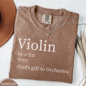 May include: Espresso-colored t-shirt with the word "Violin" in white, along with its definition. A silver necklace with a pendant rests on the shirt. The shirt also has the text "God's gift to Orchestra".