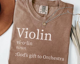 May include: Espresso-colored t-shirt with the word "Violin" in white, along with its definition. A silver necklace with a pendant rests on the shirt. The shirt also has the text "God's gift to Orchestra".