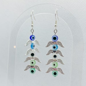 May include: A pair of dangle earrings featuring a stacked design. Each earring has a series of colorful evil eye beads in blue, white, black, and green, separated by silver-toned wing charms. The earrings hang from silver-colored hooks.