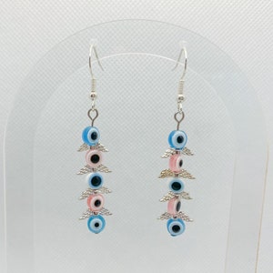 May include: A pair of dangle earrings featuring blue and pink evil eye beads with silver-colored wing accents. The earrings have silver-colored hooks and are displayed against a white background.